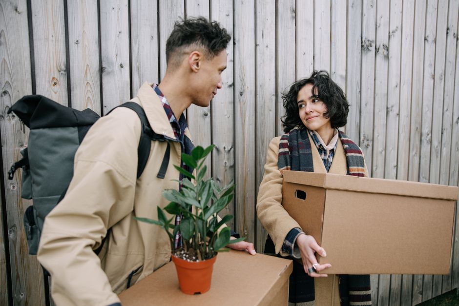 A man and a woman are standing outdoors against a wooden fence, engaged in a house relocation process. The man, on the left, is wearing a beige jacket and has a black backpack on his shoulders; he is holding a small potted plant with green leaves and a brown paper bag. The woman, on the right, has curly dark hair and is wearing a checked shirt under a camel coat, with a multicolored plaid scarf around her neck. She is holding a large cardboard box and appears to be smiling or in conversation with the man. The scene suggests an outdoor loading or unloading activity related to furniture transport and packing, typical of moving services offered by Man with Van Uxbridge. The natural lighting highlights the packing materials and the preparations involved in home relocation, with the fence providing a neutral background for the moving context.
