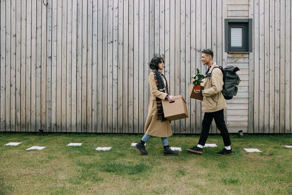 Two young individuals, a woman and a man, are walking side by side on a paved pathway adjacent to a wooden building exterior. The woman, on the left, is carrying a medium-sized cardboard box in each hand, wearing a beige coat, black boots, and a colorful scarf around her neck. She has short, dark curly hair and appears to be engaged in conversation with the man. The man, on the right, is carrying a small potted plant in a brown cardboard box and has a black backpack on his shoulders. He is dressed in a beige jacket, black trousers, and black shoes, with short, dark hair styled neatly. Both are smiling and looking at each other as they walk. The background features vertical wooden cladding, a small window with a black frame and closed shutters, and the pavement is lined with evenly spaced stone slabs on a well-maintained grassy area, indicating movement as part of a house relocation or furniture transport process, coordinated by [COMPANY_NAME] for a house removals service.