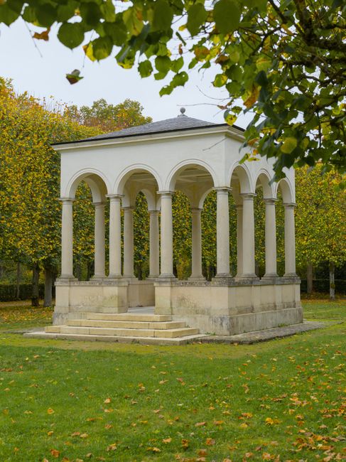 A wooden gazebo with a pitched roof, constructed from light-colored timber, is situated on a grassy area surrounded by tall, mature trees with green foliage, casting shadows on the ground. The gazebo 