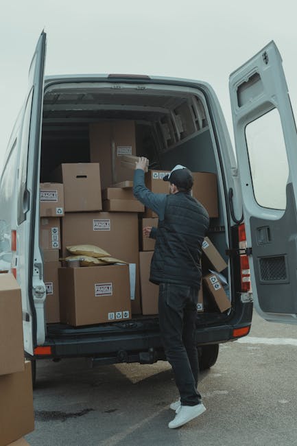 A person wearing a dark jacket, grey pants, and a dark cap is loading a cardboard box into the rear of a white moving van, which is parked on an outdoor driveway. The van’s rear doors are wide open,