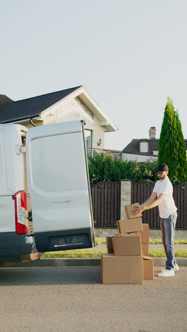 A person wearing a dark jacket, grey pants, and a dark cap is loading a cardboard box into the rear of a white moving van, which is parked on an outdoor driveway. The van’s rear doors are wide open,